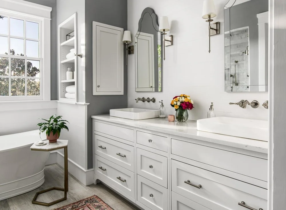 Bright modern bathroom with white cabinetry, vessel sinks, and a large mirror. Features a window, a plant on a side table, and a flower vase on the counter.