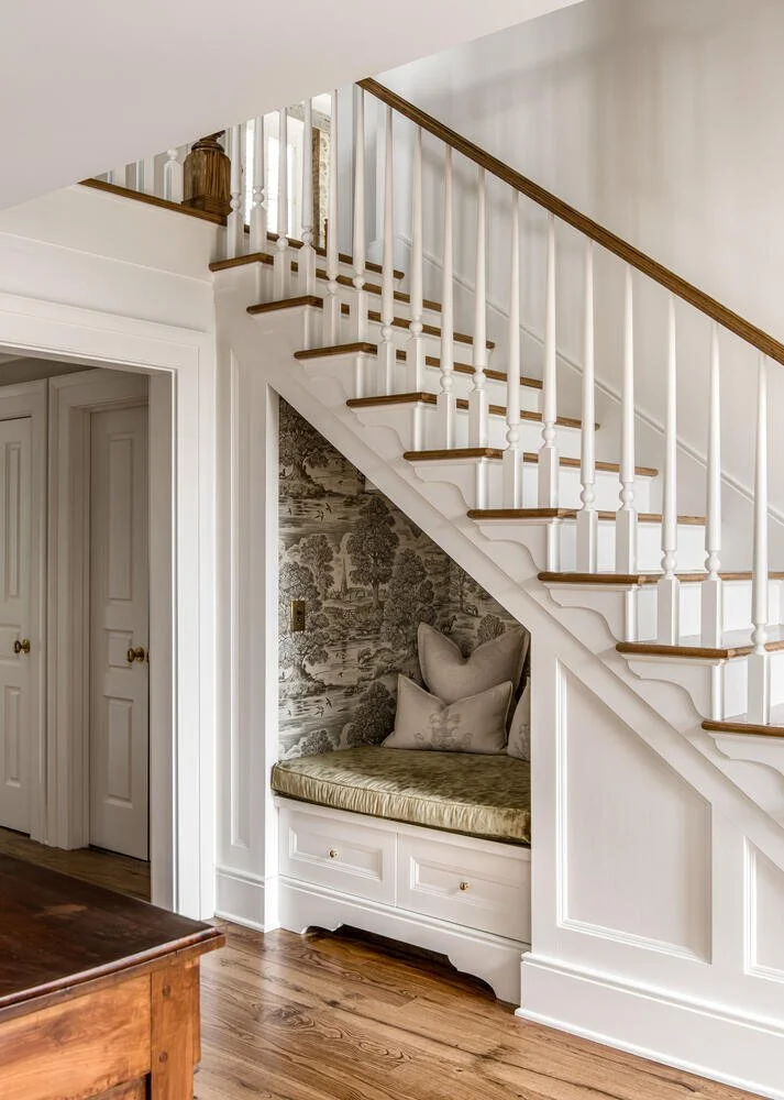 Interior of a home showing a staircase with white risers and wooden treads, a built-in bench with pillows beneath the stairs, patterned wallpaper behind the bench, and white trim and walls.