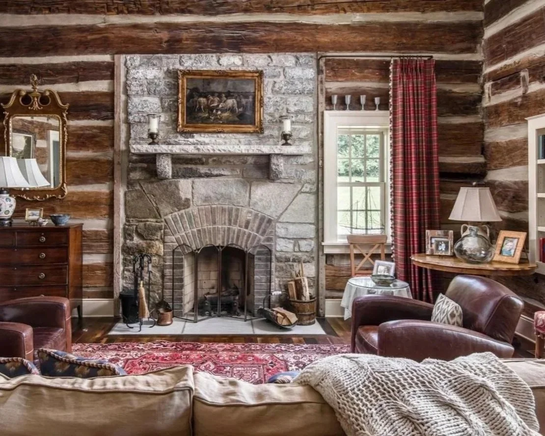 A cozy living room with a stone fireplace, wooden walls, and vintage furniture. There's a sofa with a knitted blanket, armchairs, a rug, a side table with a lamp, and picture frames.