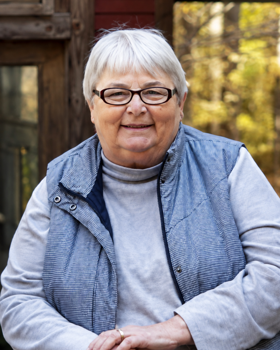 A smiling elderly woman with short white hair, wearing glasses, a gray sweater, and a checkered blue vest, sitting outdoors with autumn leaves in the background.