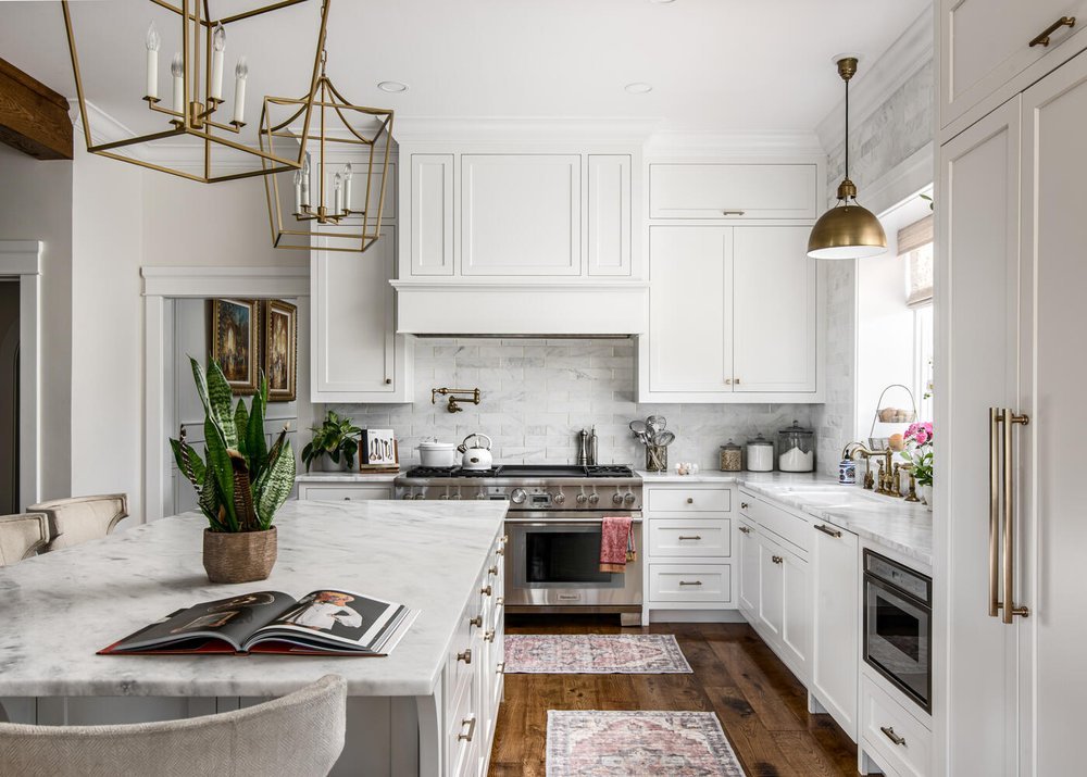 Modern kitchen with white cabinets, marble countertops, stainless steel appliances, and gold accents. Includes a large island with a potted plant and open magazine, pendant lighting, and hardwood floors.
