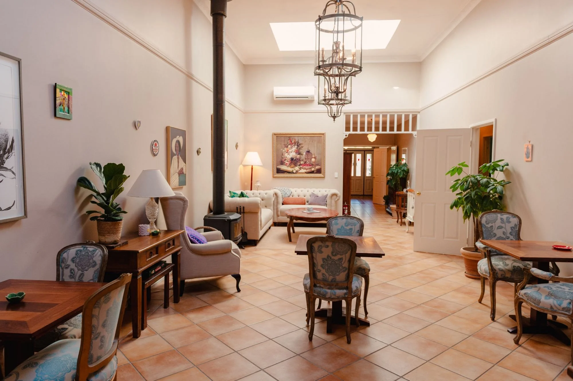 Spacious living room with a fireplace, a chandelier, and several chairs and tables. There are decorative paintings on the walls, potted plants, and a door to another room.