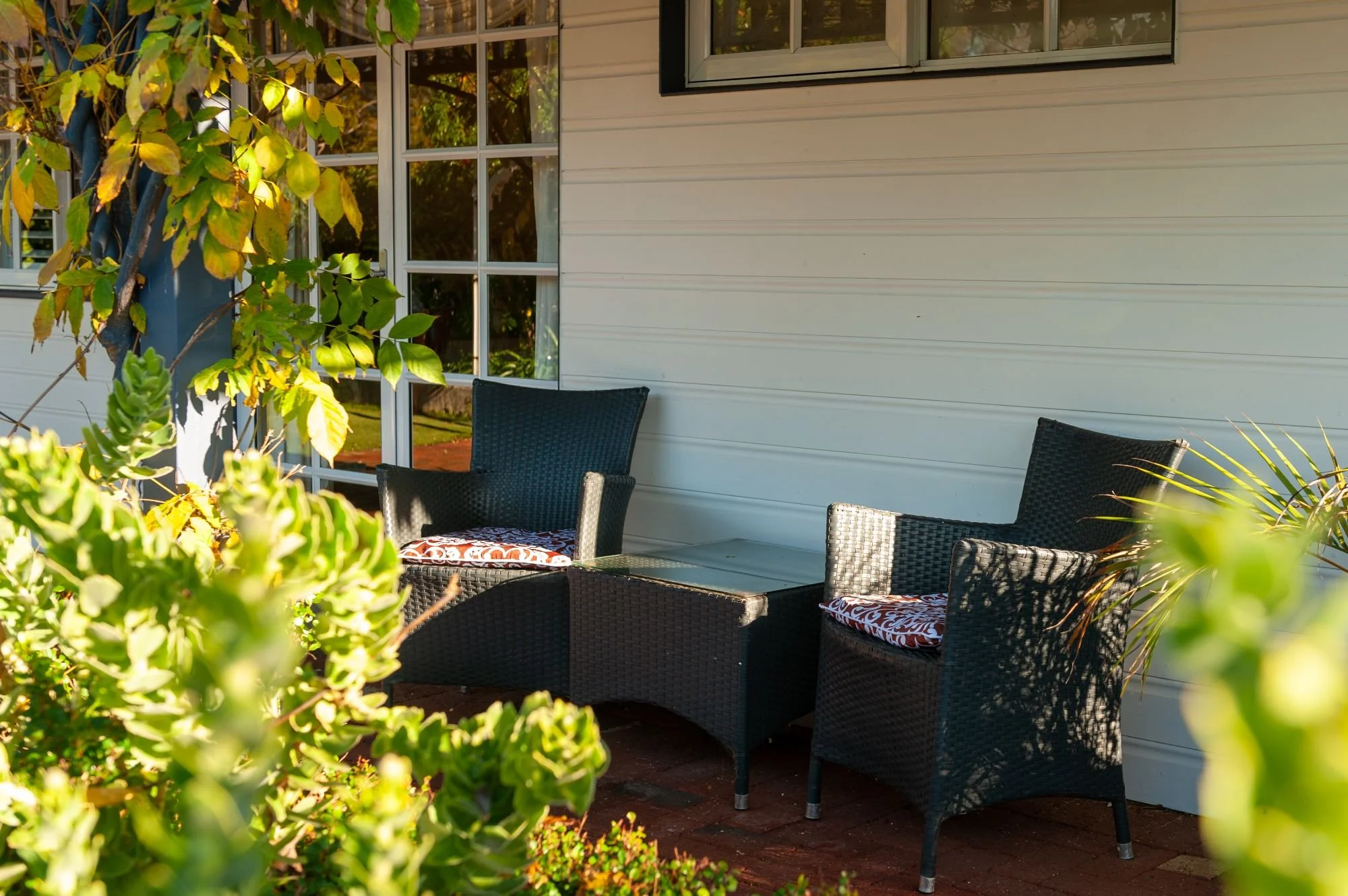 Outdoor patio with black wicker chairs with patterned cushions, surrounded by greenery.