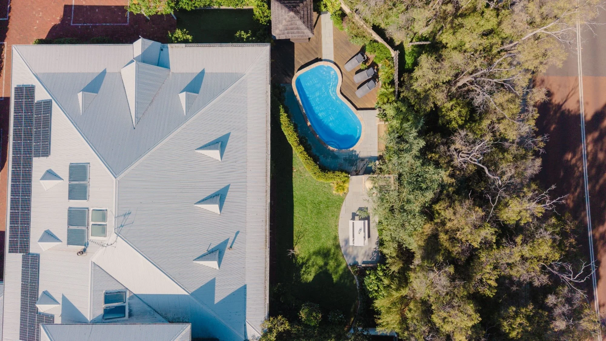 Aerial view of Lanterns Retreat. Next to the house is a backyard with a swimming pool, surrounded by a wooden deck, lounge chairs, and a private garden lined by trees.