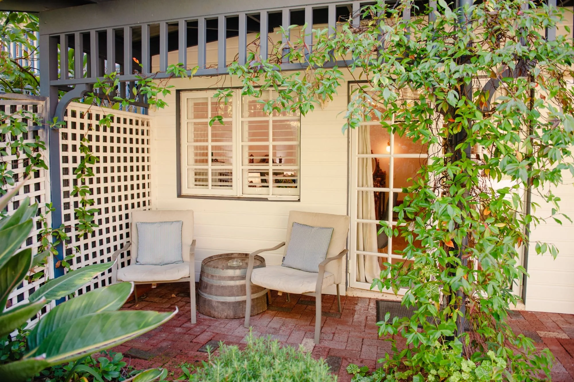 A cozy outdoor patio with two white cushioned chairs, a small round wooden table, surrounded by green foliage and a trellis, with a house with white siding and large windows in the background.