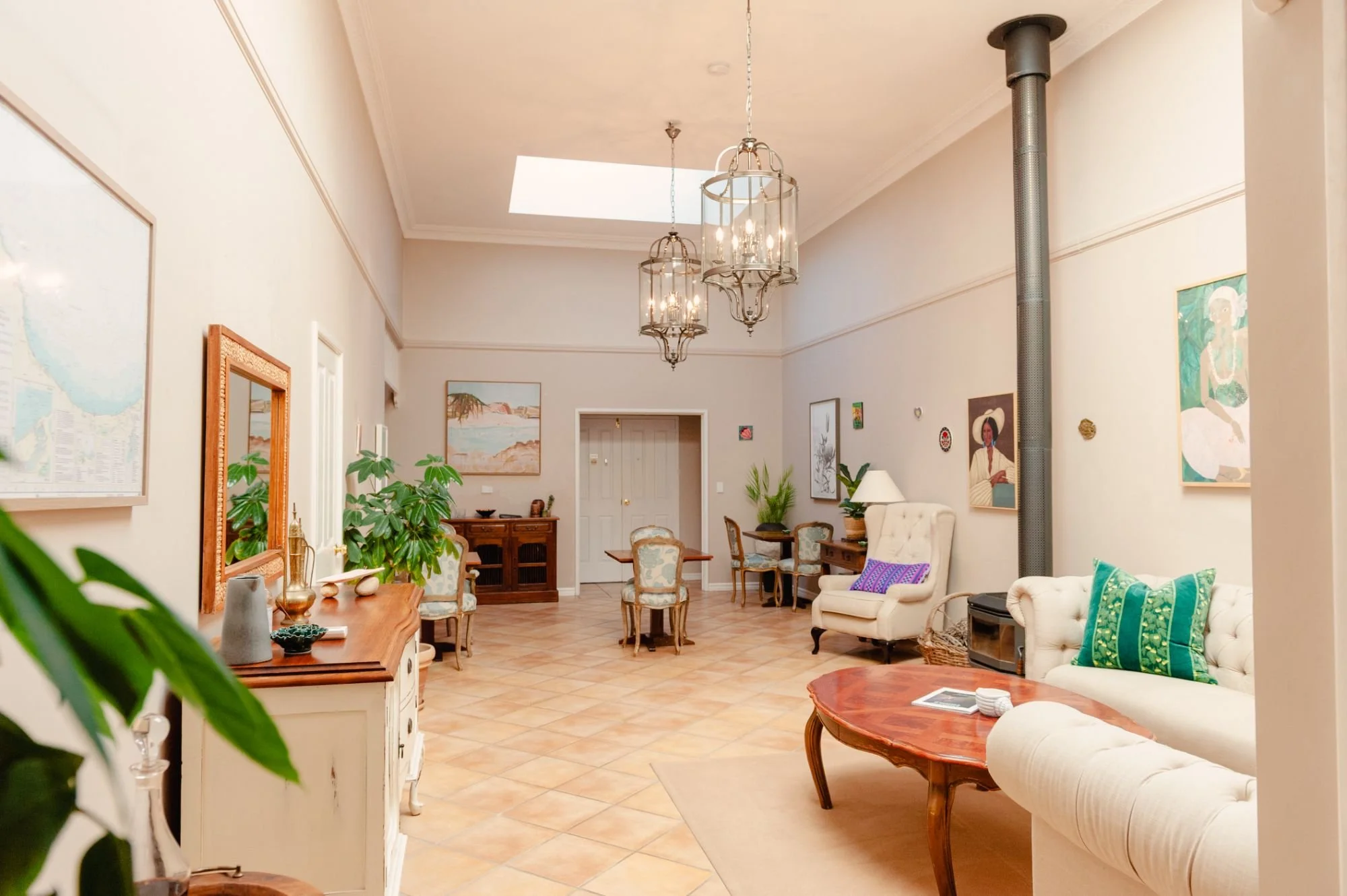 Spacious living room with light-colored walls, ceiling, and tiled floor. Features multiple paintings, a chandelier, a white sofa with colorful pillows, a round wooden coffee table, and several chairs, with a skylight and side cabinets.