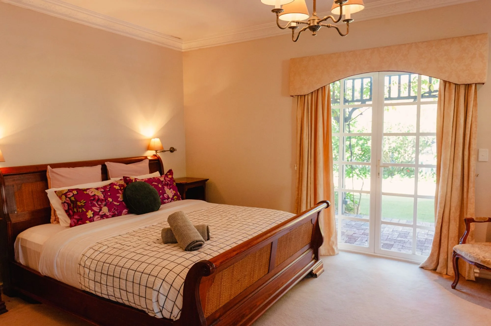 A bedroom with a large wooden bed, decorative pillows, rolled towels, and a window with curtains and a view of greenery outside.