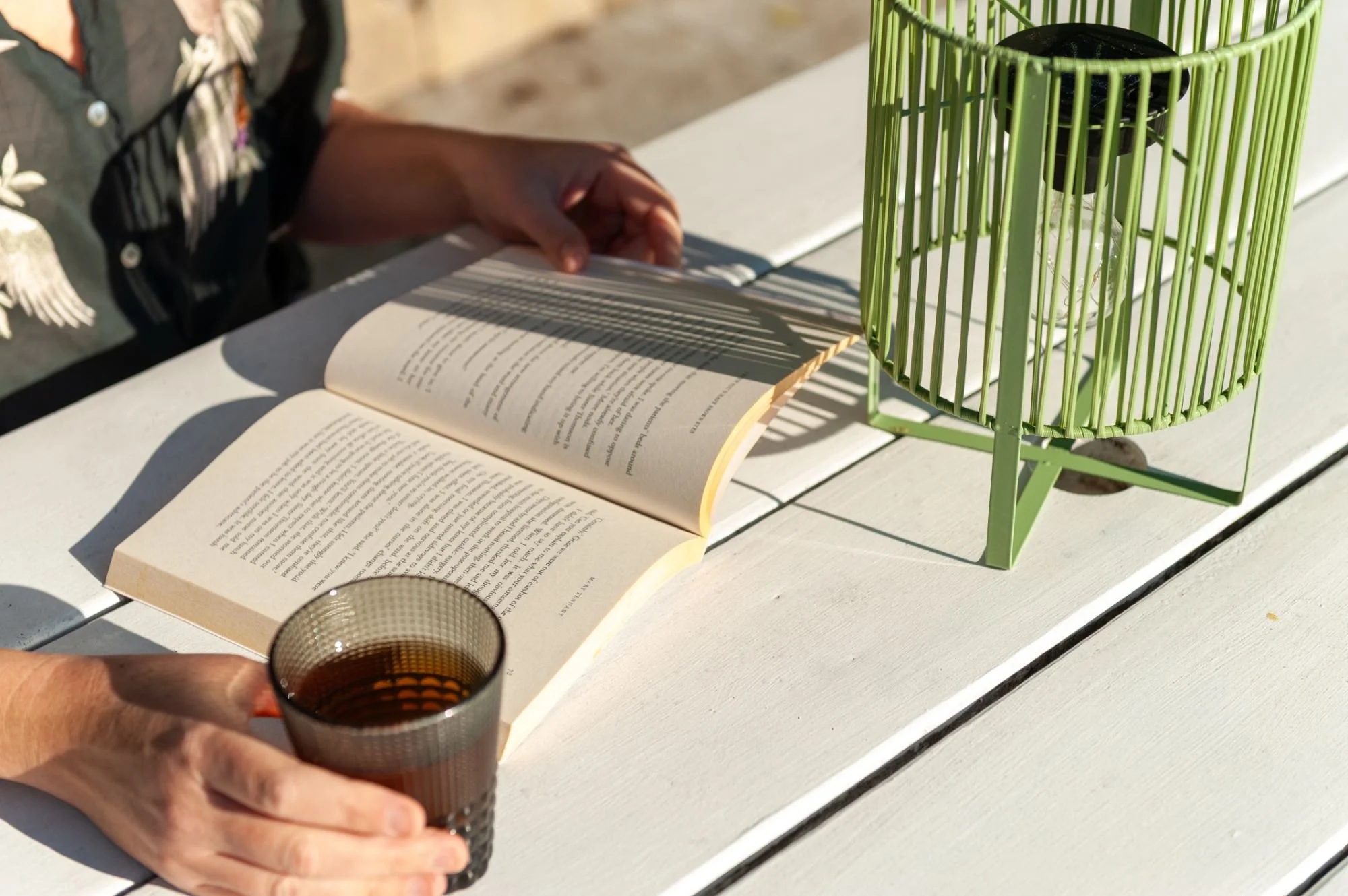 Person holding a glass of dark beverage over an open book on a table outdoors, with a green decorative lantern nearby.