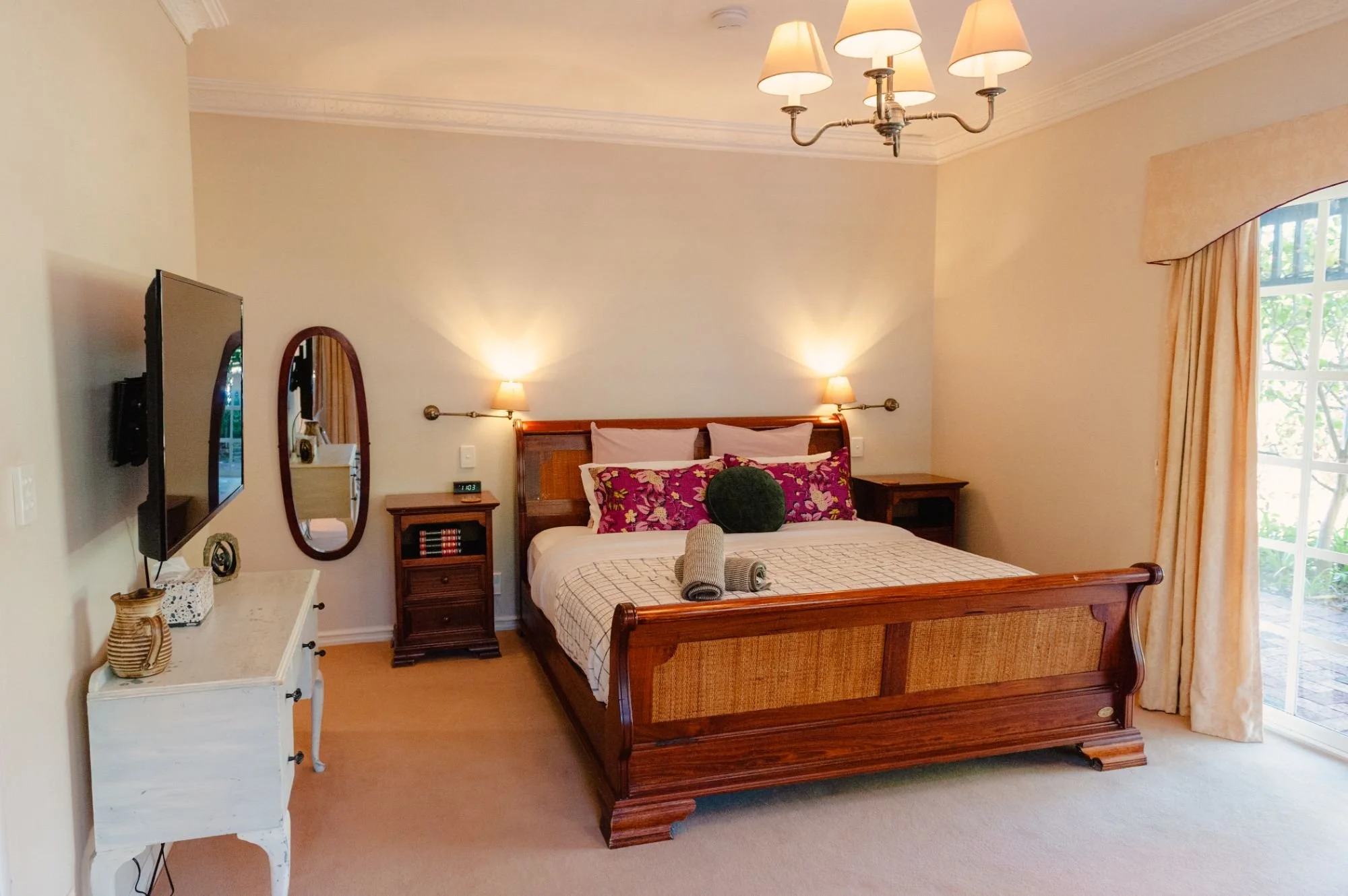 A bedroom with a wooden bed, nightstands, and lamps. The bed has pink and floral pillows with gray towels on it. A window with beige curtains is on the right, and a flat-screen TV is on the left. The room has cream-colored walls and a chandelier with five lamps hanging from the ceiling.