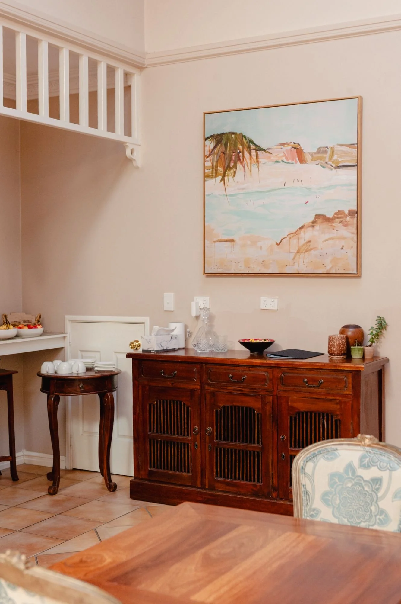 A cozy dining room with a wooden sideboard, a small table with cups and a bowl, a painting of a beach scene, and a patterned armchair.