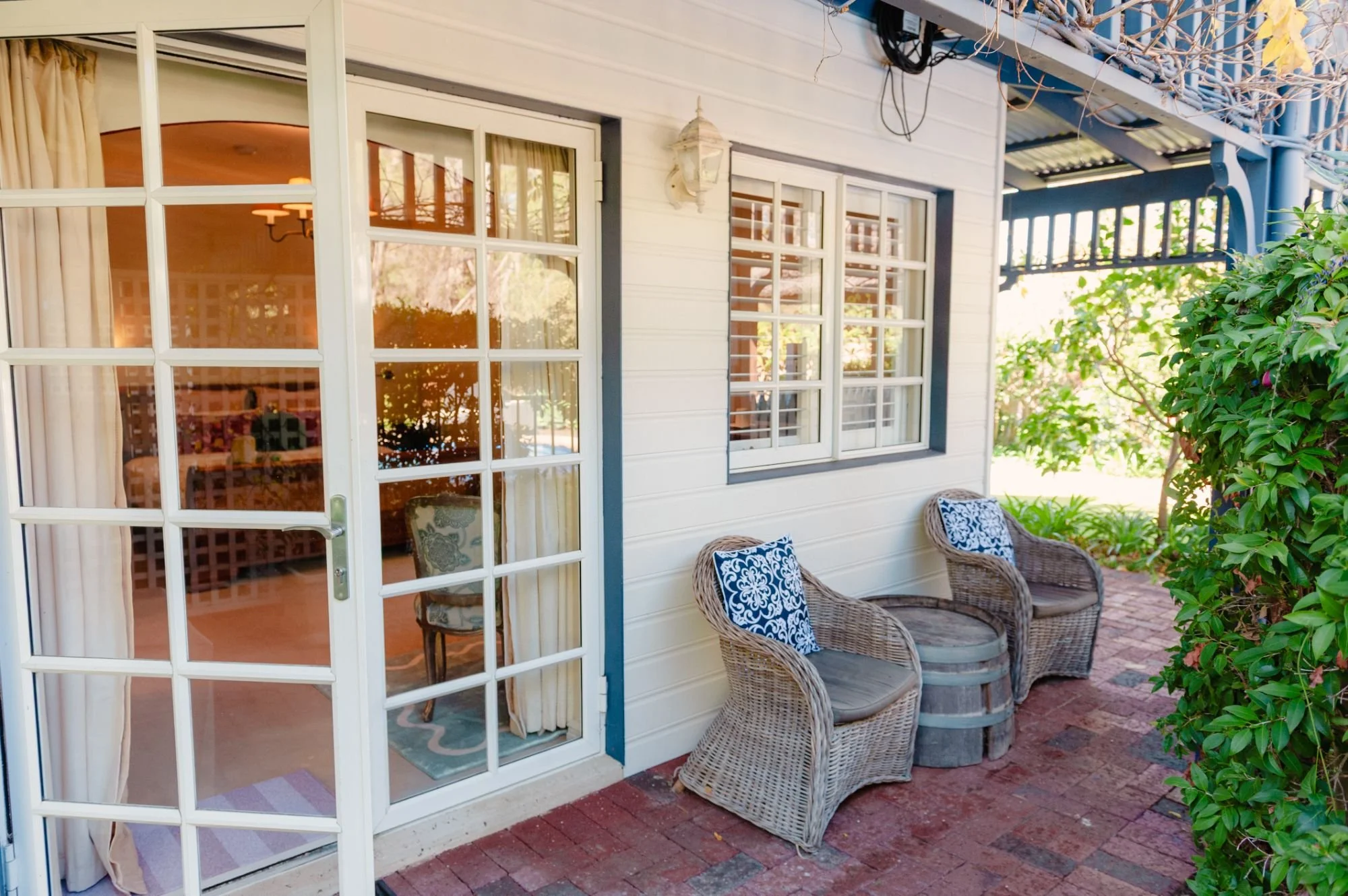 Exterior view of a porch with a white door with glass panes, two wicker chairs with patterned cushions, and a green bush on the right side. The porch has a brick floor and is partially covered with a pergola.