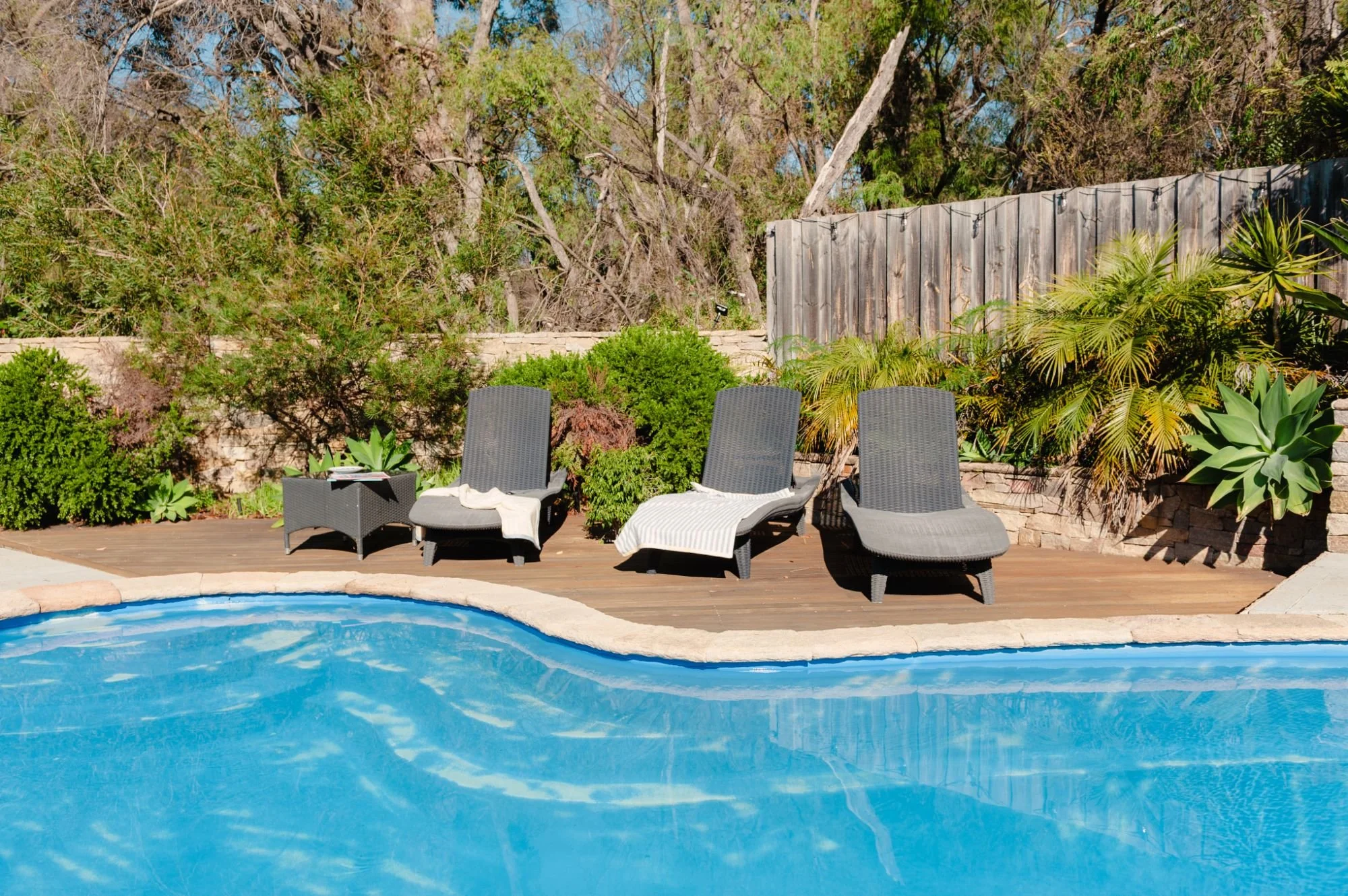 Lanterns Retreat swimming pool, three black lounge chairs, and lush green plants and trees behind a wooden fence.