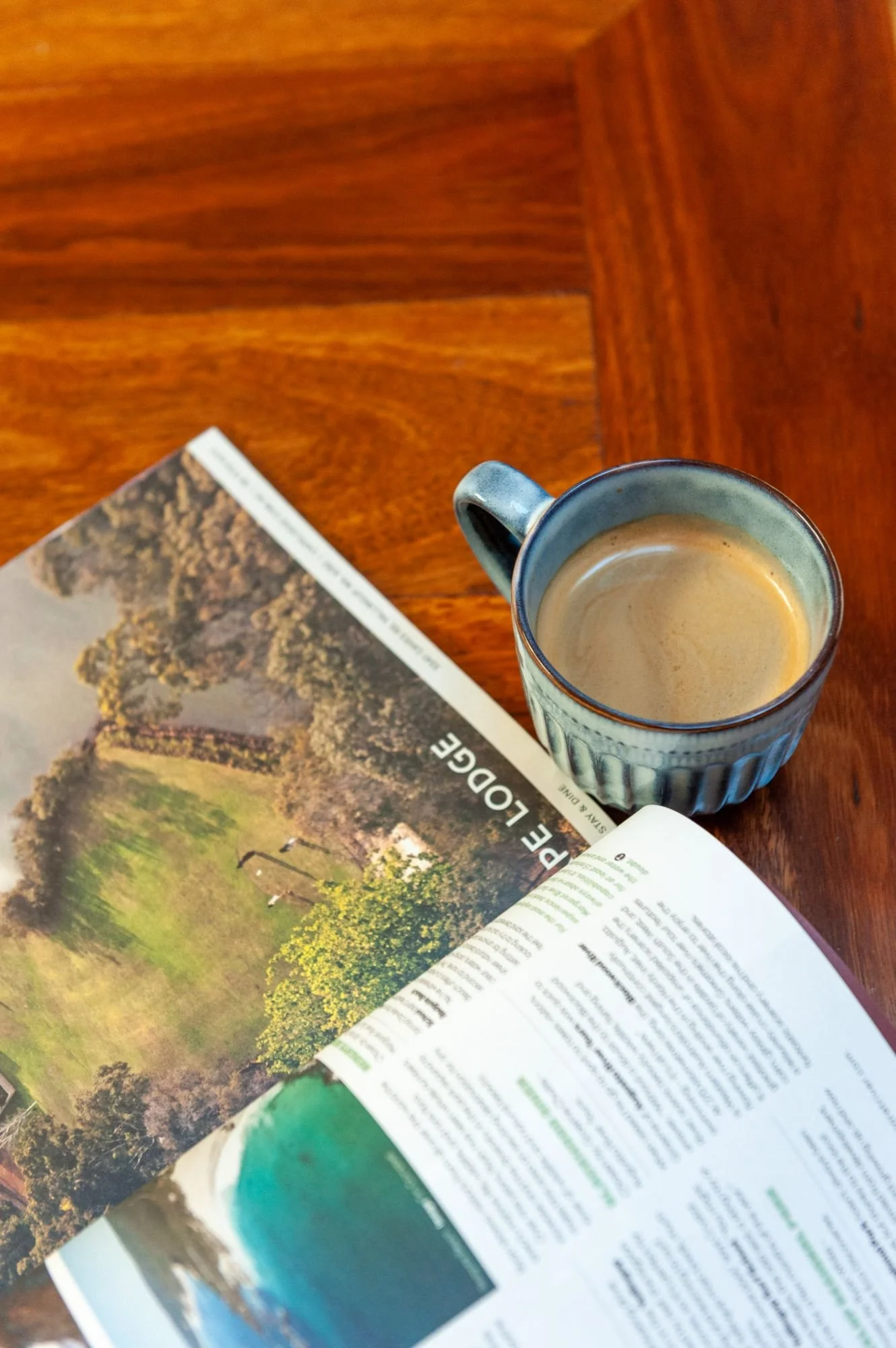 A mug of coffee or tea on a wooden table next to an open magazine or travel guide.