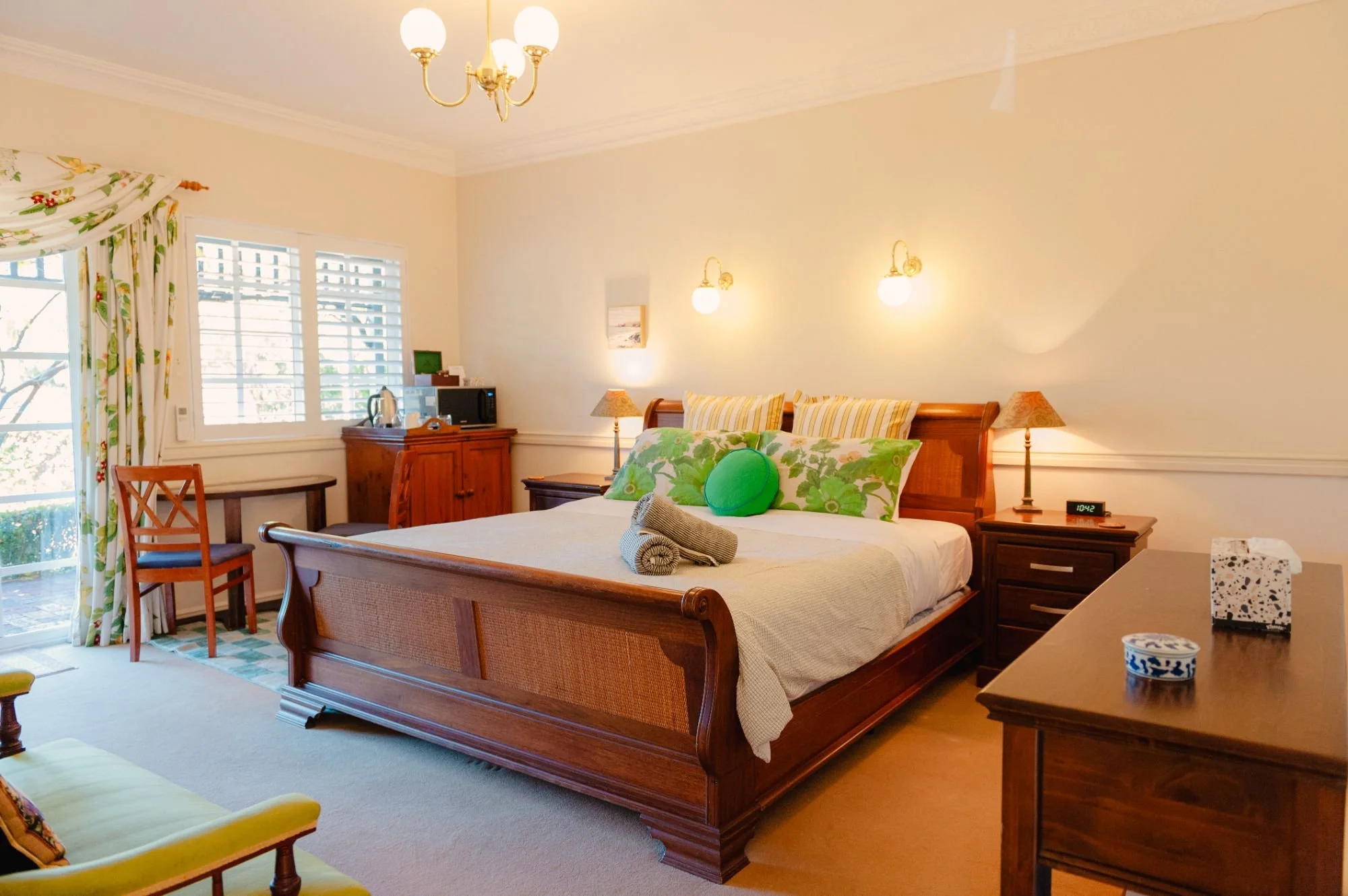 A bedroom with a wooden bed, white bedding, green accent pillows, and a rolled towel. There are nightstands with lamps, a window with floral curtains, and a small desk with a microwave and kettle. The room has a chandelier and wall-mounted lights.