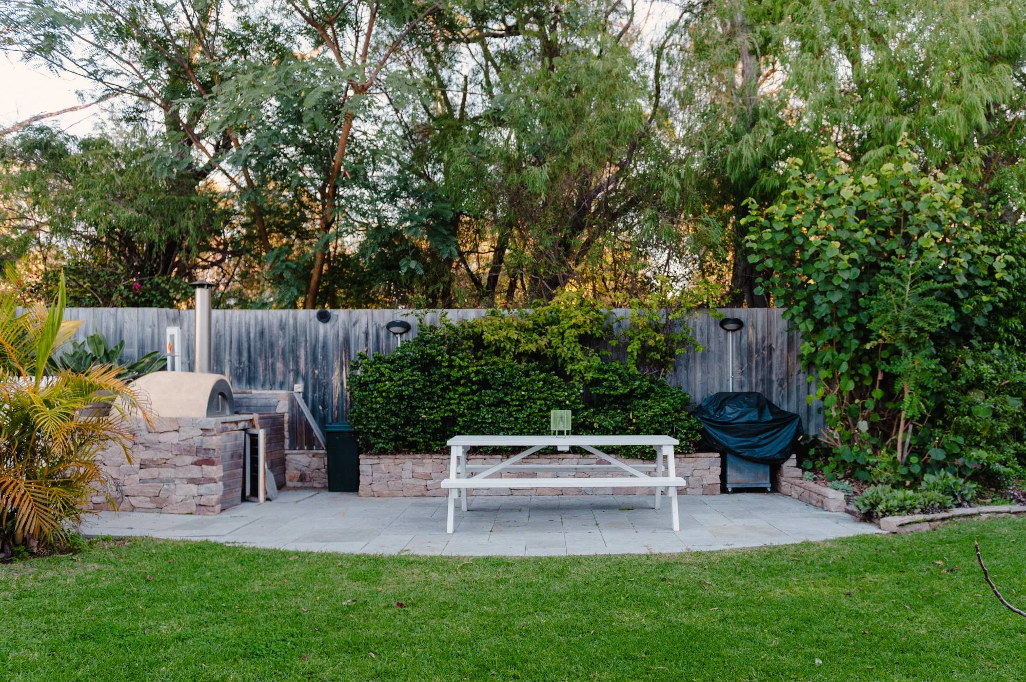 Backyard patio area with a lawn, stone and wood fencing, trees, and plants, featuring a white picnic table and an outdoor grill.