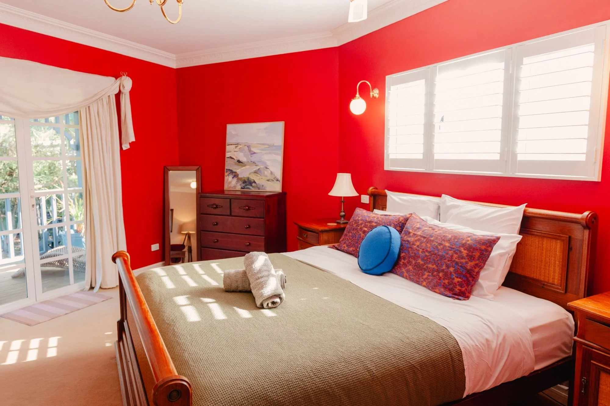A bedroom with red walls, white window shutters, a wooden bed with white bedding and colorful pillows, a bedside table with a lamp, and a dresser with a painting and a mirror.