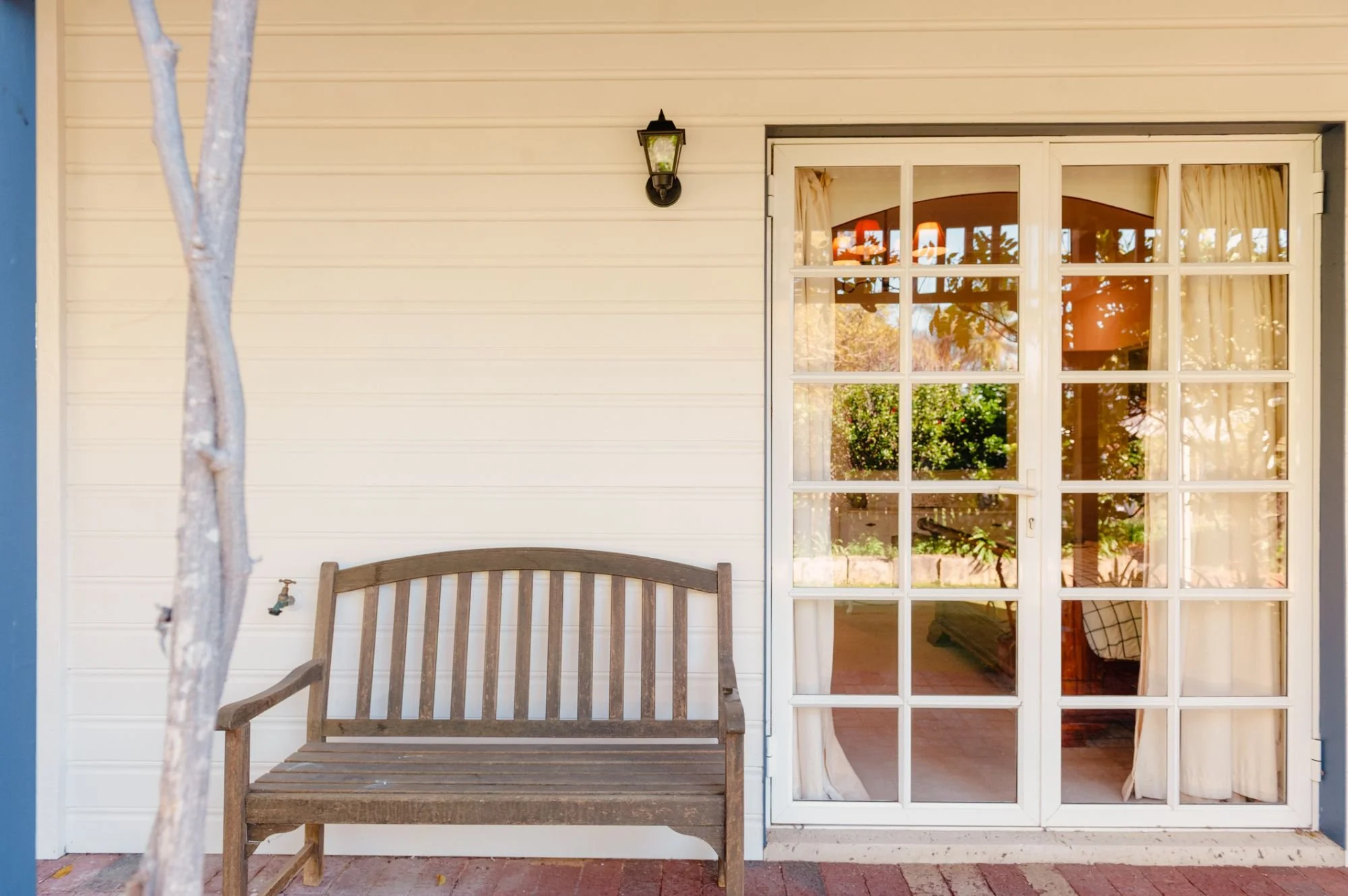 A porch area with a wooden bench against a beige wall, a black outdoor light fixture, and glass sliding doors with curtains, revealing a view of a garden inside.