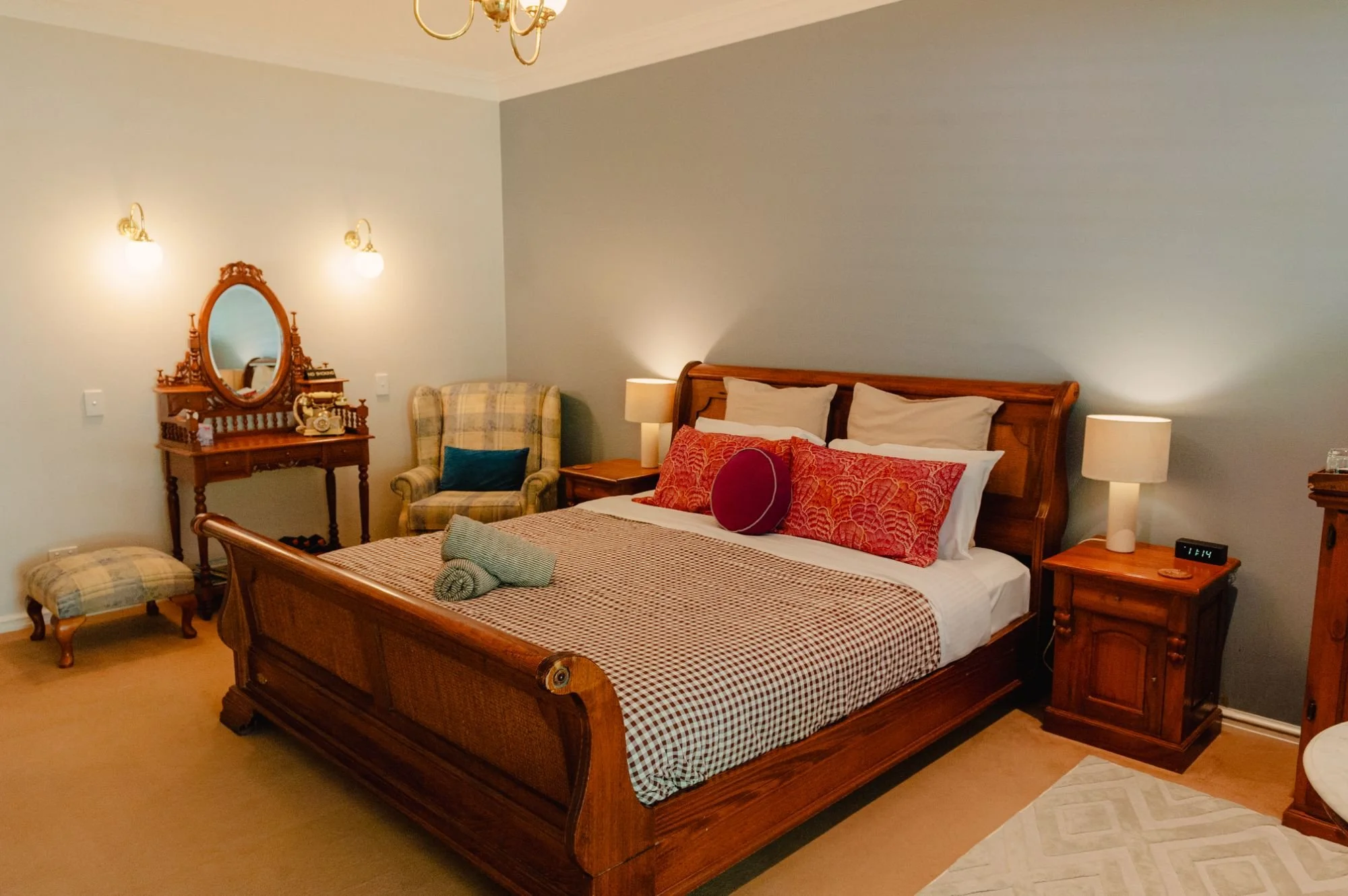 A bedroom with a wooden bed frame, bedspread, pillows, and towels. There are matching wooden nightstands with lamps, an upholstered chair, a dresser with a mirror, and a chandelier. Soft lighting creates a cozy atmosphere.