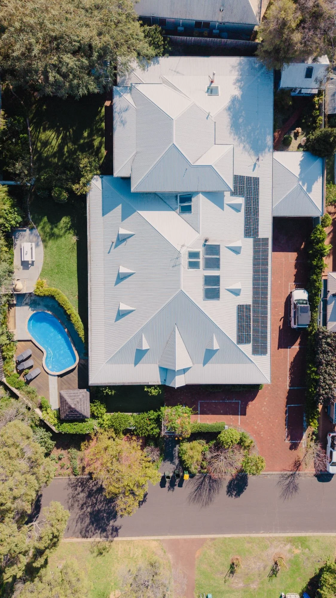 An aerial view the Lanterns Retreat house with a metal roof, solar panels, and surrounding yard, trees, and a swimming pool.