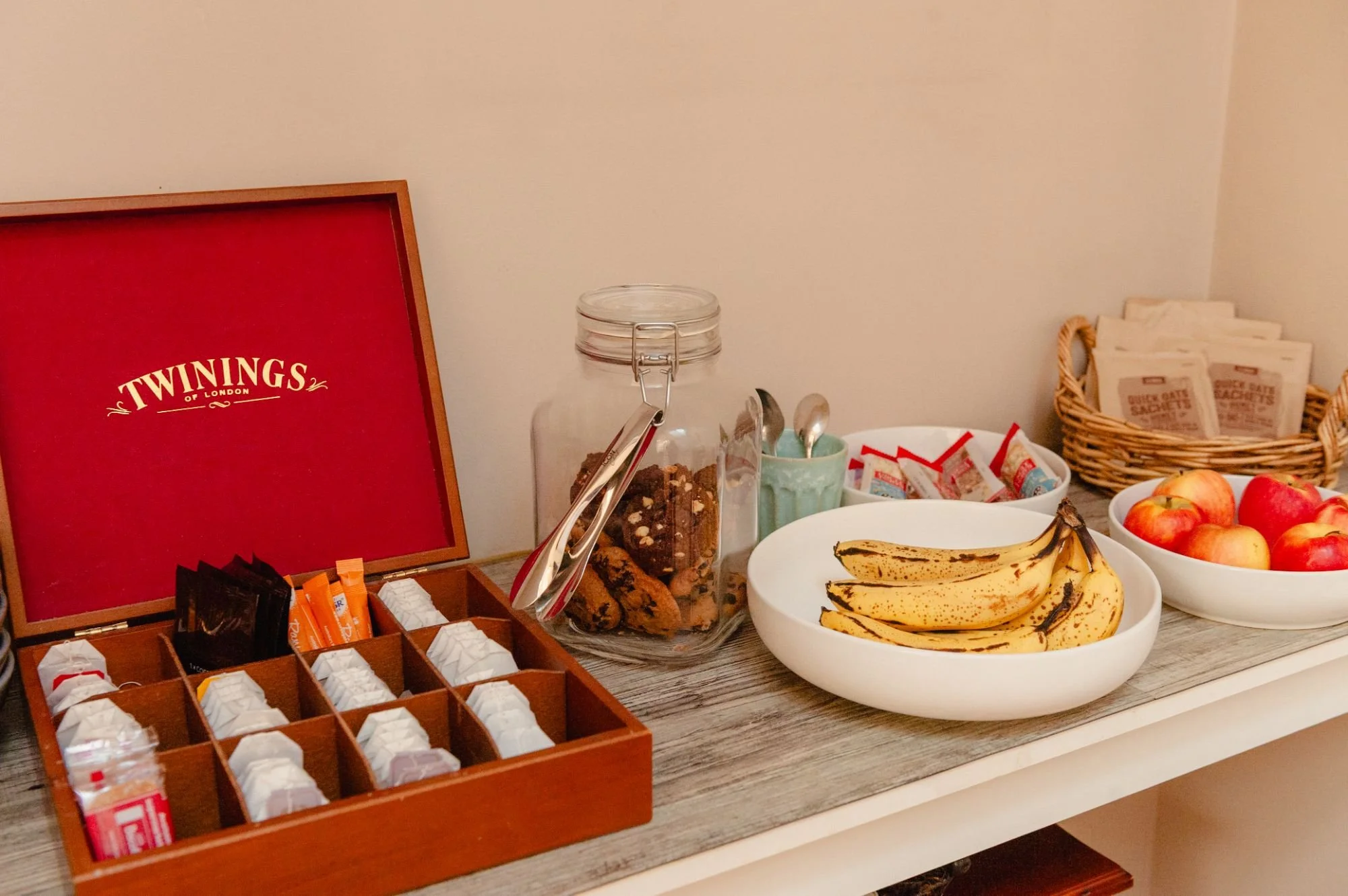 Assorted snacks and fruits on a table, including a box of Twinings tea, a jar of cookies, bananas, apples, and various packets and cups.