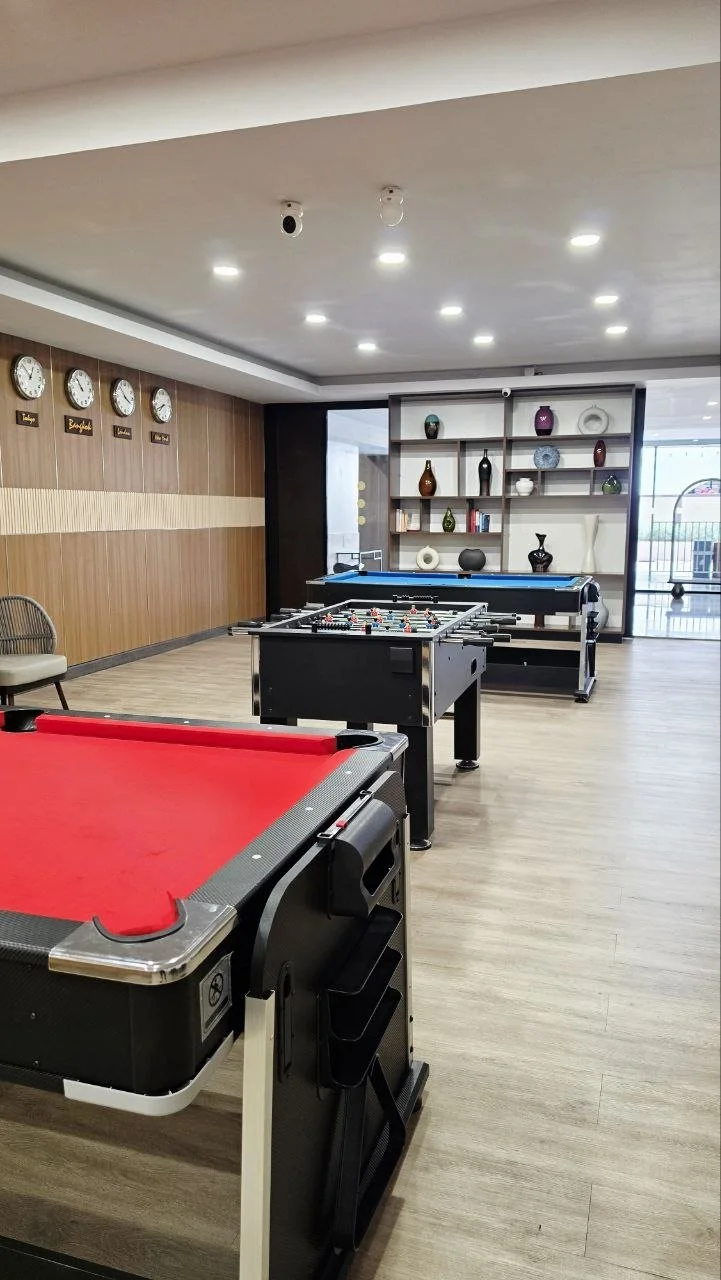 An indoor game room with a red pool table, a foosball table, and a blue snooker table. Light-colored wood flooring, wall clocks, and decorative shelves with vases and books on the back wall. Large windows letting in natural light.