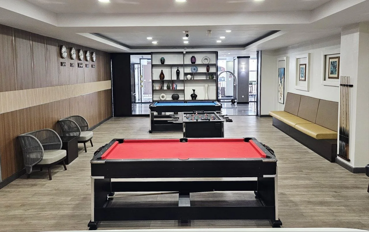 A game room with a red pool table in the foreground, a black and blue foosball table in the middle, and a blue or black corner billiards table in the background. The room has light-colored wood floors, a seating area with three chairs on the left, a 