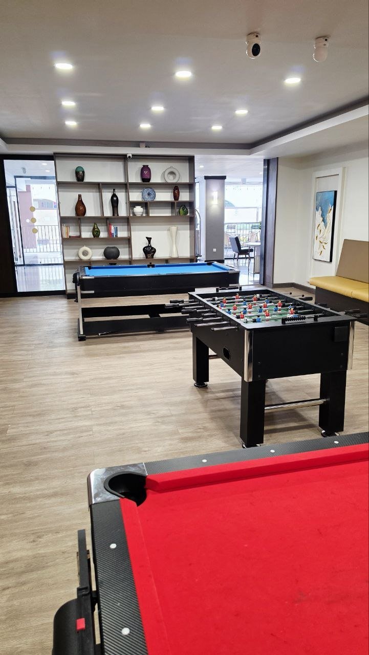 An indoor game room with a red pool table in the foreground, a black foosball table in the middle, and a blue pool table in the background. There are shelves with decorative items and a seating area near the window.