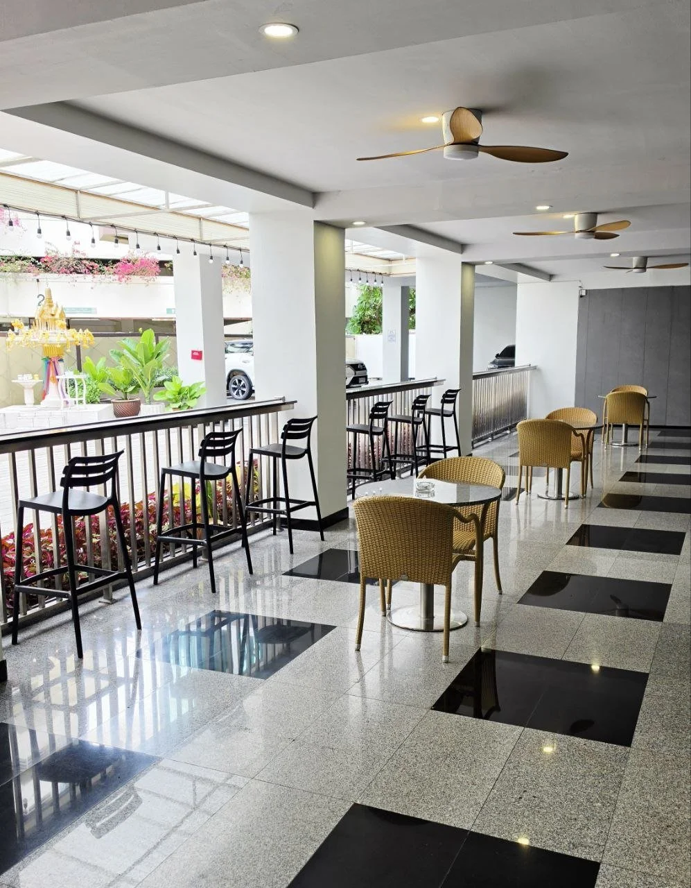 Interior view of a bright, modern cafe or restaurant with black and beige wicker chairs, some round tables, a long bar with black chairs, ceiling fans, and large windows with planters and outdoor view.
