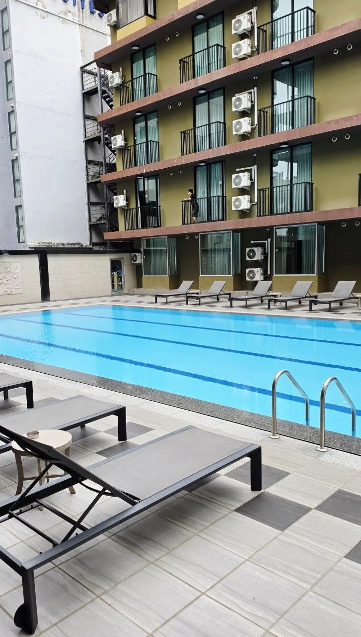 An outdoor hotel pool area with lounge chairs and a building with balconies overlooking the pool.