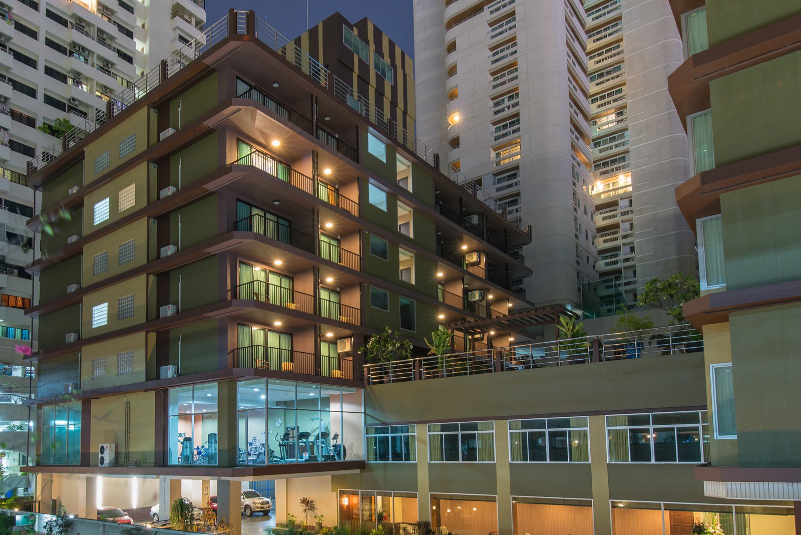 Night view of a modern apartment building with multiple floors featuring balconies, some with potted plants. The ground floor has a fitness center with exercise equipment visible through large windows and underground parking with cars. The building is illuminated with exterior lights, and neighboring high-rise buildings are visible in the background.