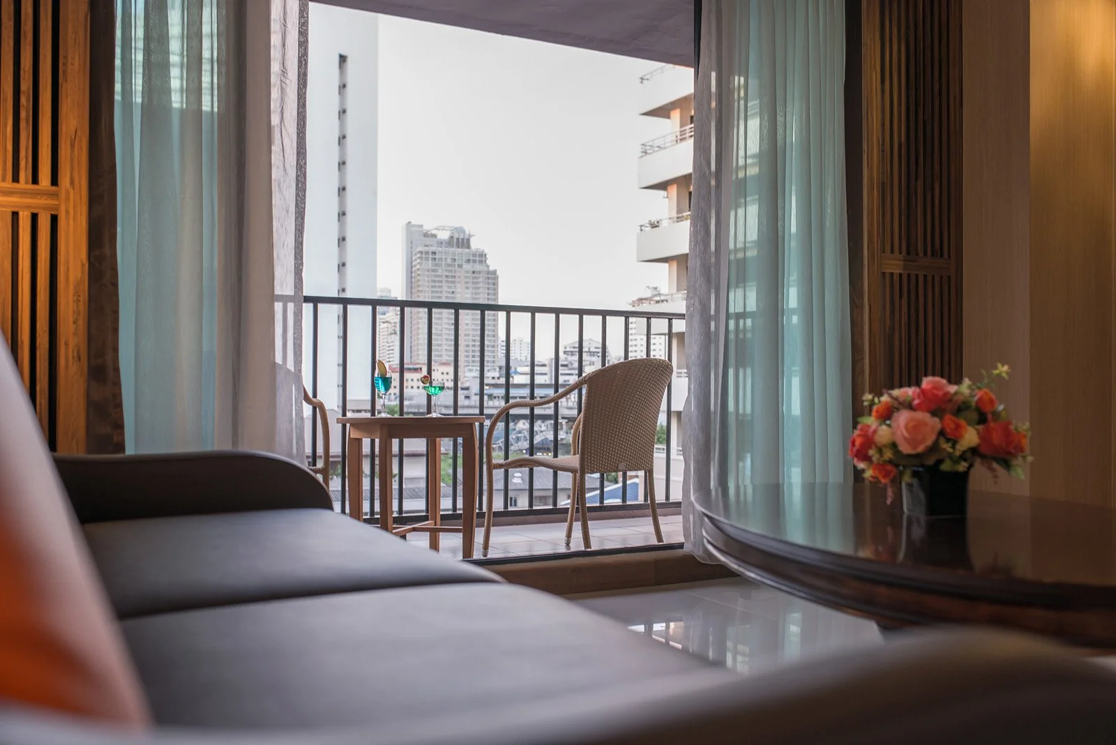 Hotel room with a balcony outside view of city buildings, a small table with colorful drinks, chairs, a sofa, a round table with a flower bouquet, and sheer curtains.