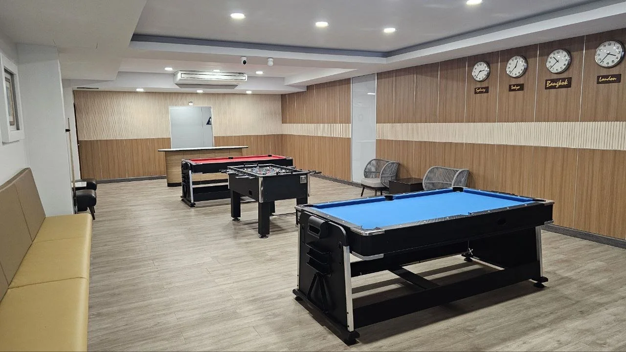 An empty game room with two pool tables, one with a blue felt surface and another with a red felt surface, set up for billiards. The room has wood-paneled walls, a beige bench on the left, two chairs on the right, and four clocks displaying different