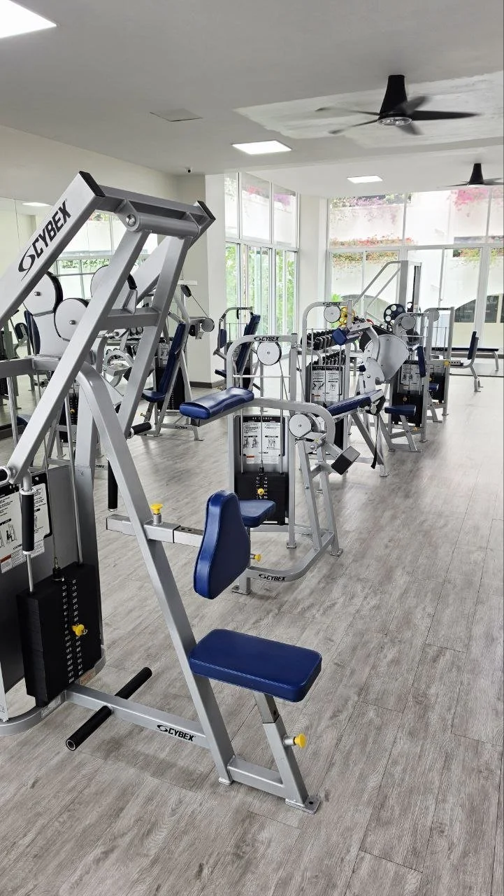 Interior of a gym with various exercise machines, large windows allowing natural light, and ceiling fans.