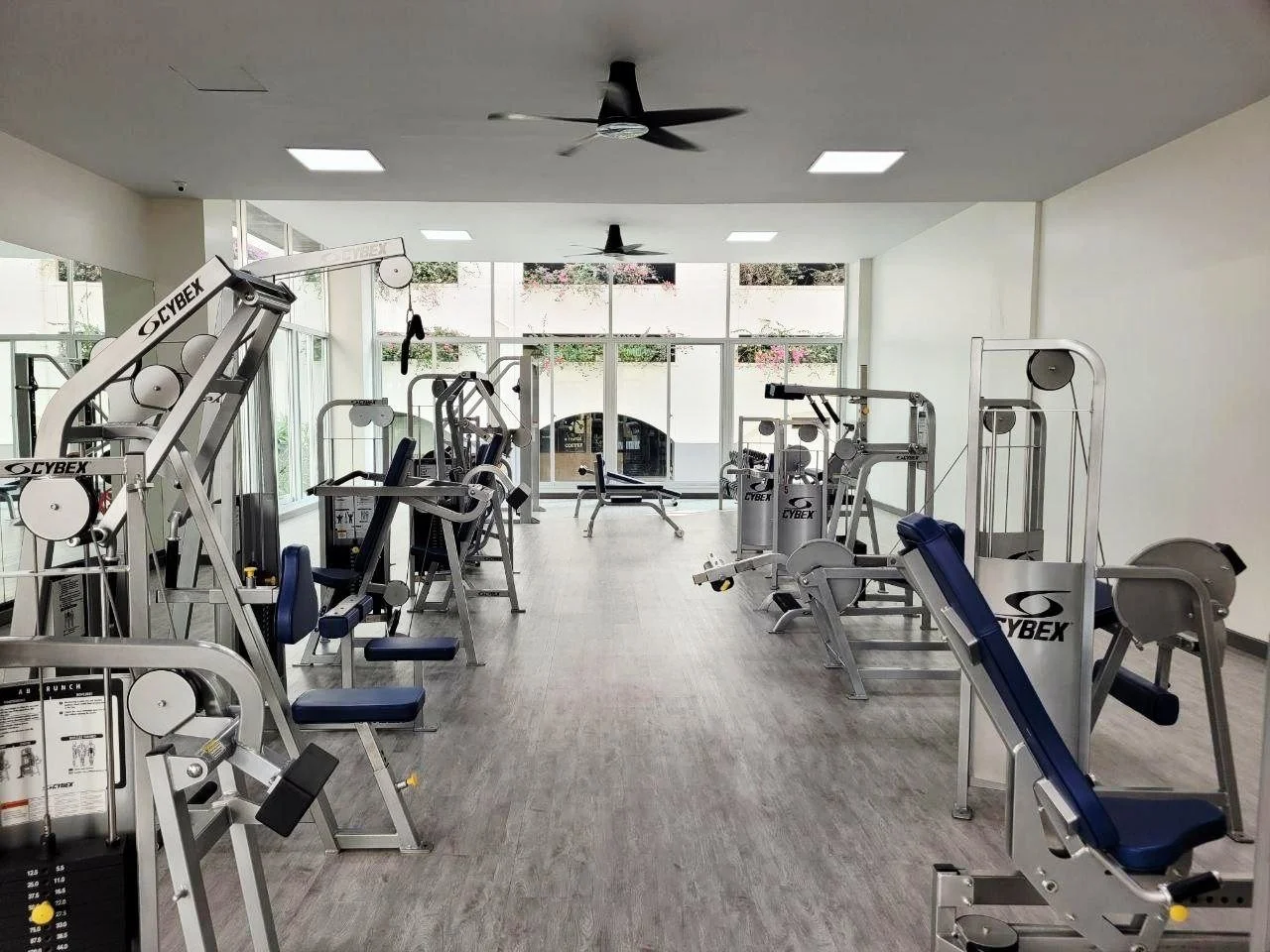 Empty gym room with various exercise machines, large windows at the back, ceiling fans, and bright lighting.