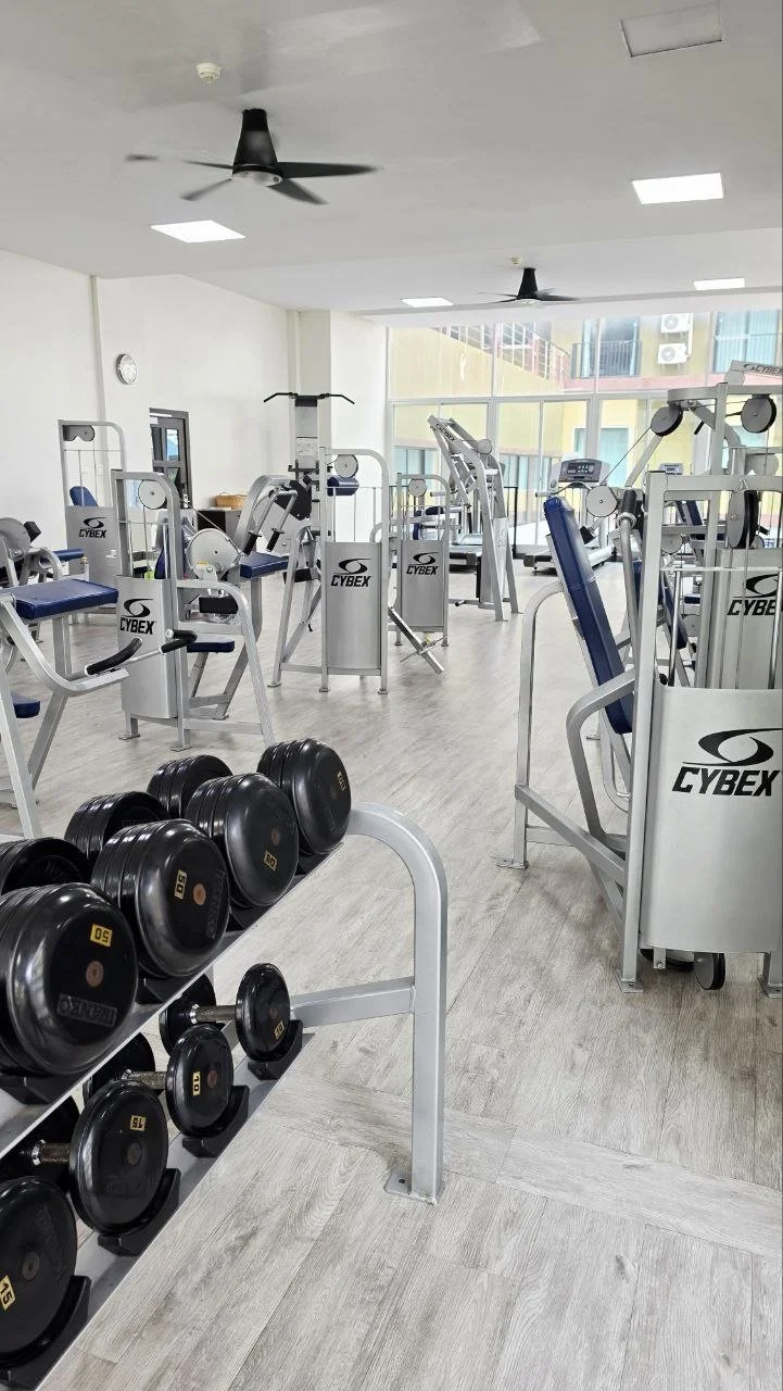Empty gym with free weights and various exercise machines, large windows at the back, ceiling fans, and a light-colored wood floor.