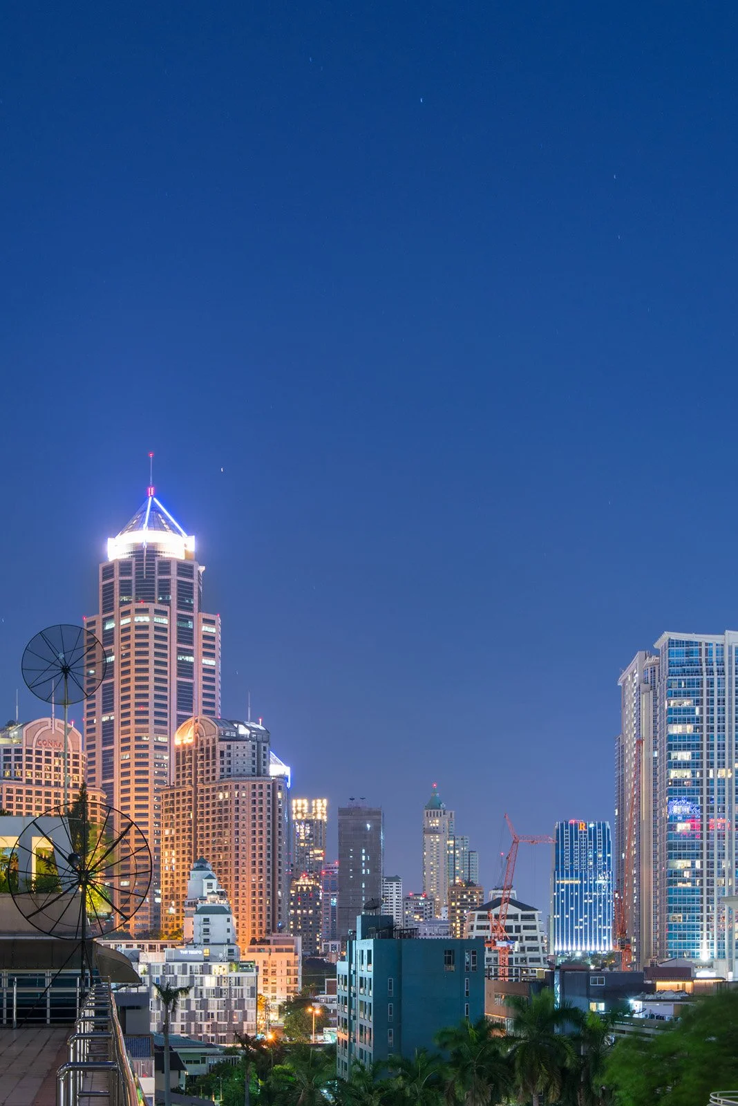 Nighttime city skyline with illuminated skyscrapers and antennas, blue sky, and some trees in the foreground.