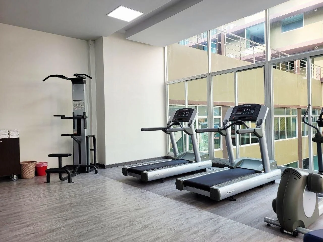 Fitness room with two treadmills near large windows, a vertical exercise machine, a small table with towels, a basket, and a red bucket.