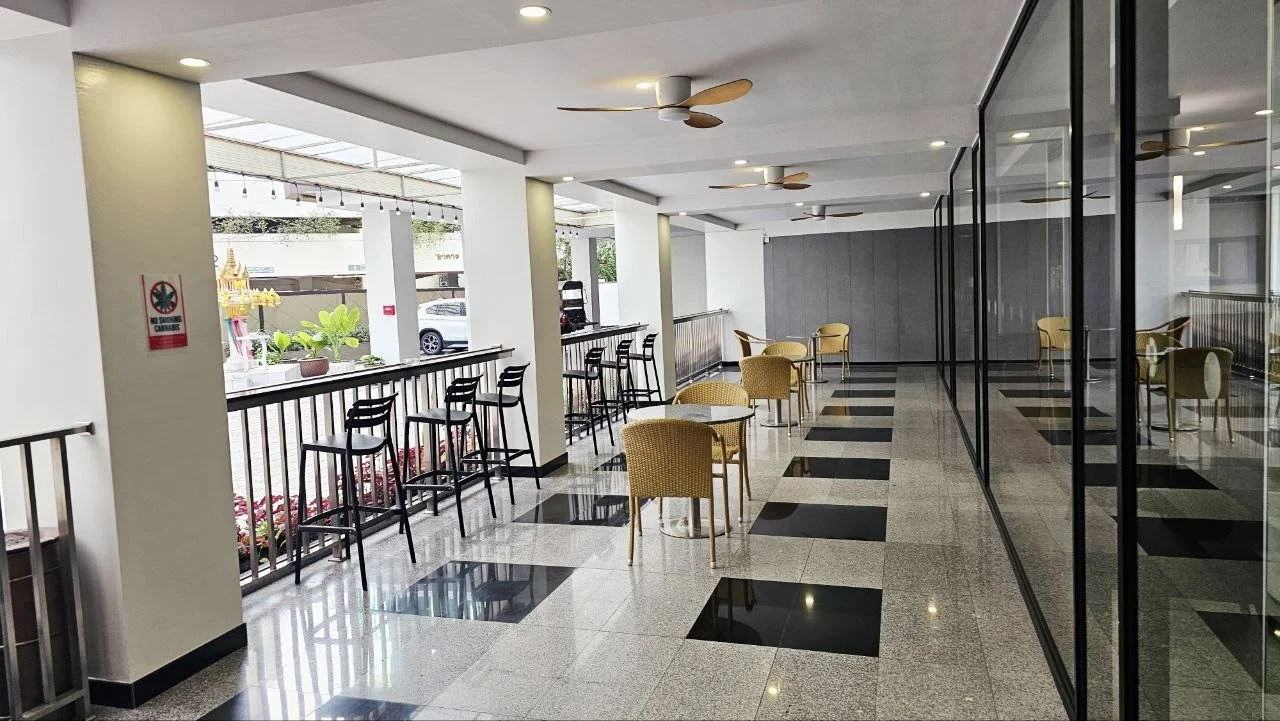 Empty indoor lounge area with yellow chairs and high tables, glass wall on the right, ceiling fans, and a balcony with black chairs outside.