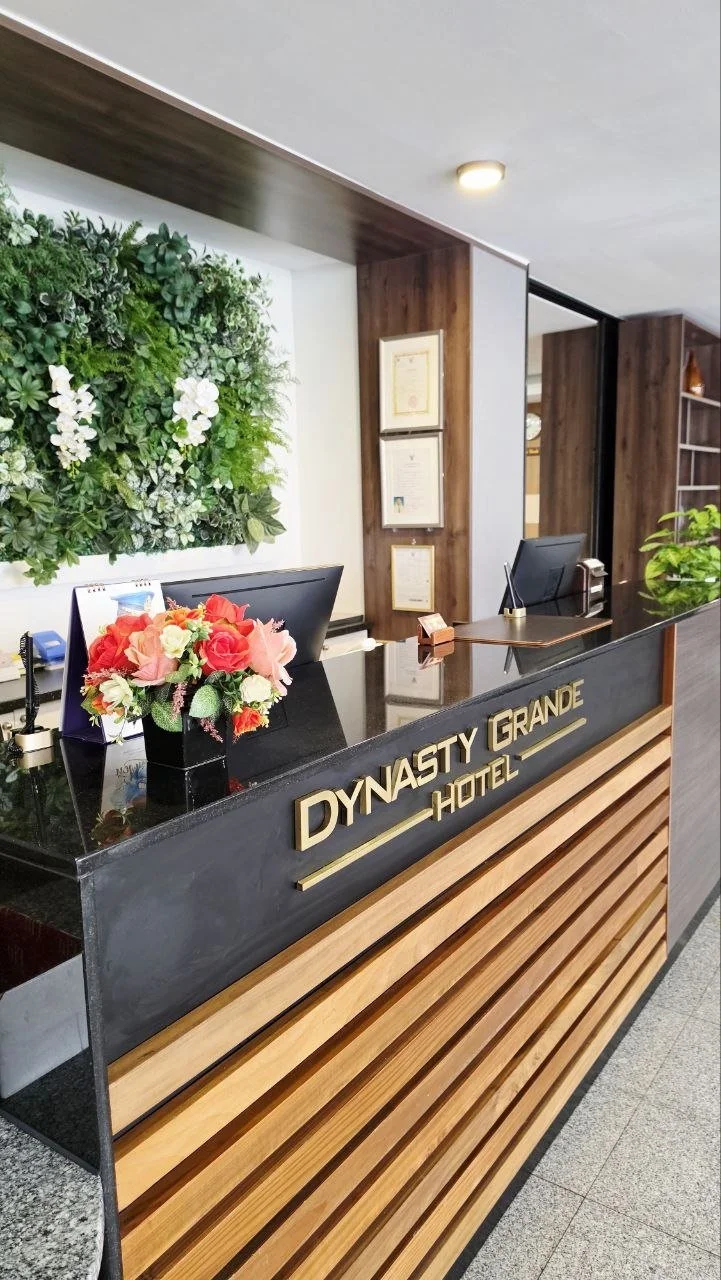 Hotel reception desk with a sign reading 'Dynasty Grande Hotel', decorated with a bouquet of flowers, green wall with white orchids, and framed certificates on a wooden wall.
