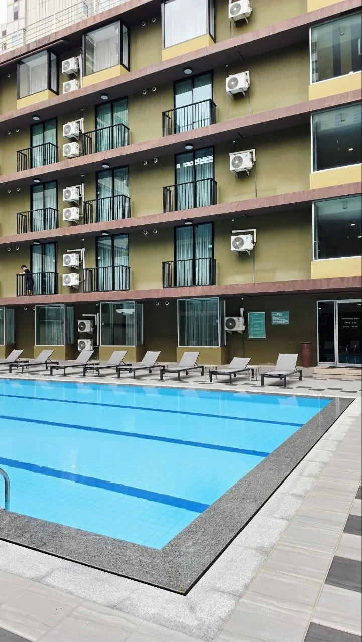View of a swimming pool at a hotel or apartment complex with lounge chairs along the deck, adjacent to a building with multiple floors featuring balconies with air conditioning units.