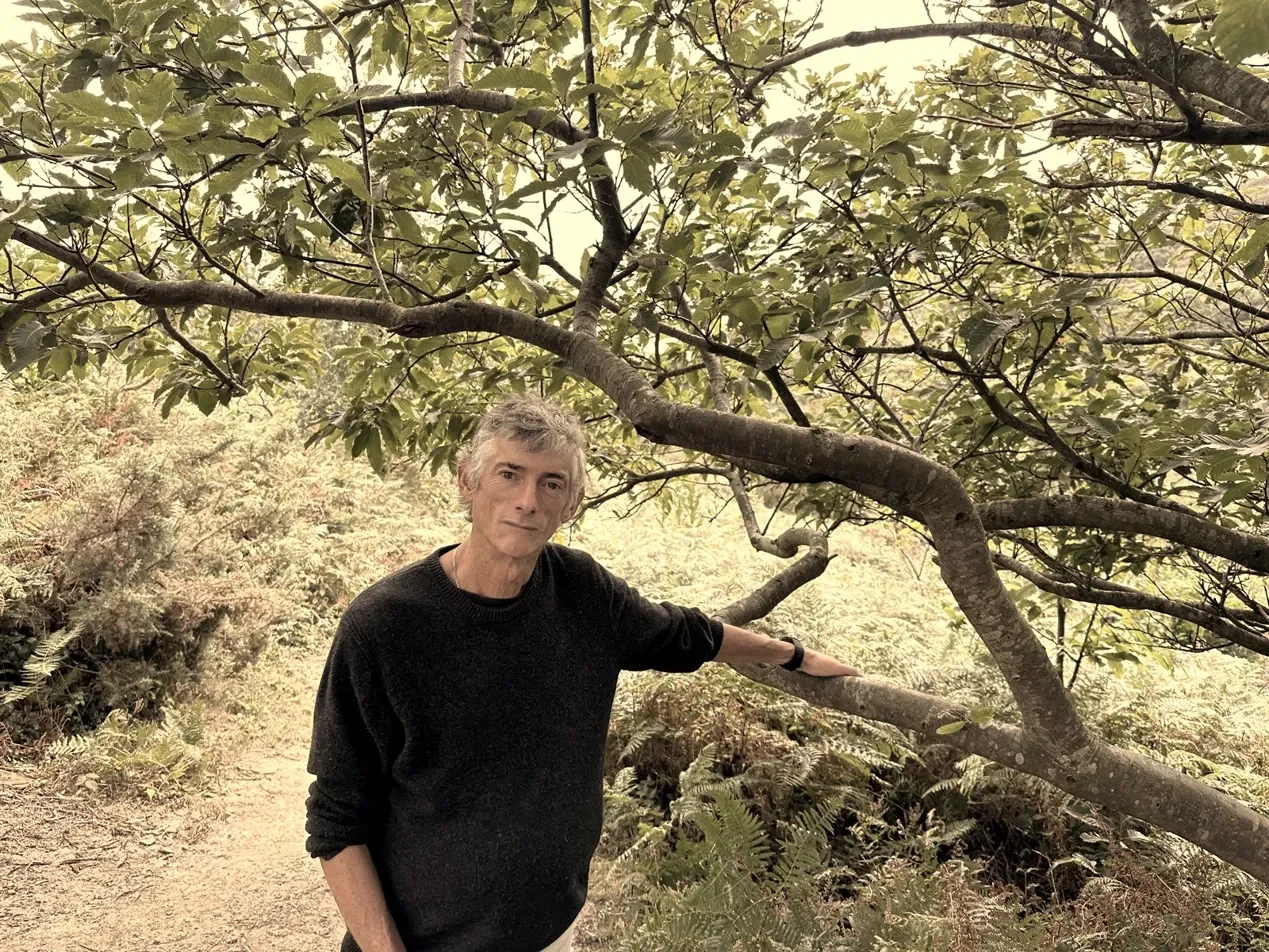 Simon Goodrich, bioresonance therapist based in Fairlight, Sussex, standing outdoors on a woodland. A middle-aged man with grey hair wearing a black sweater and light pants standing under a tree in a forest, touching a branch with his left hand.