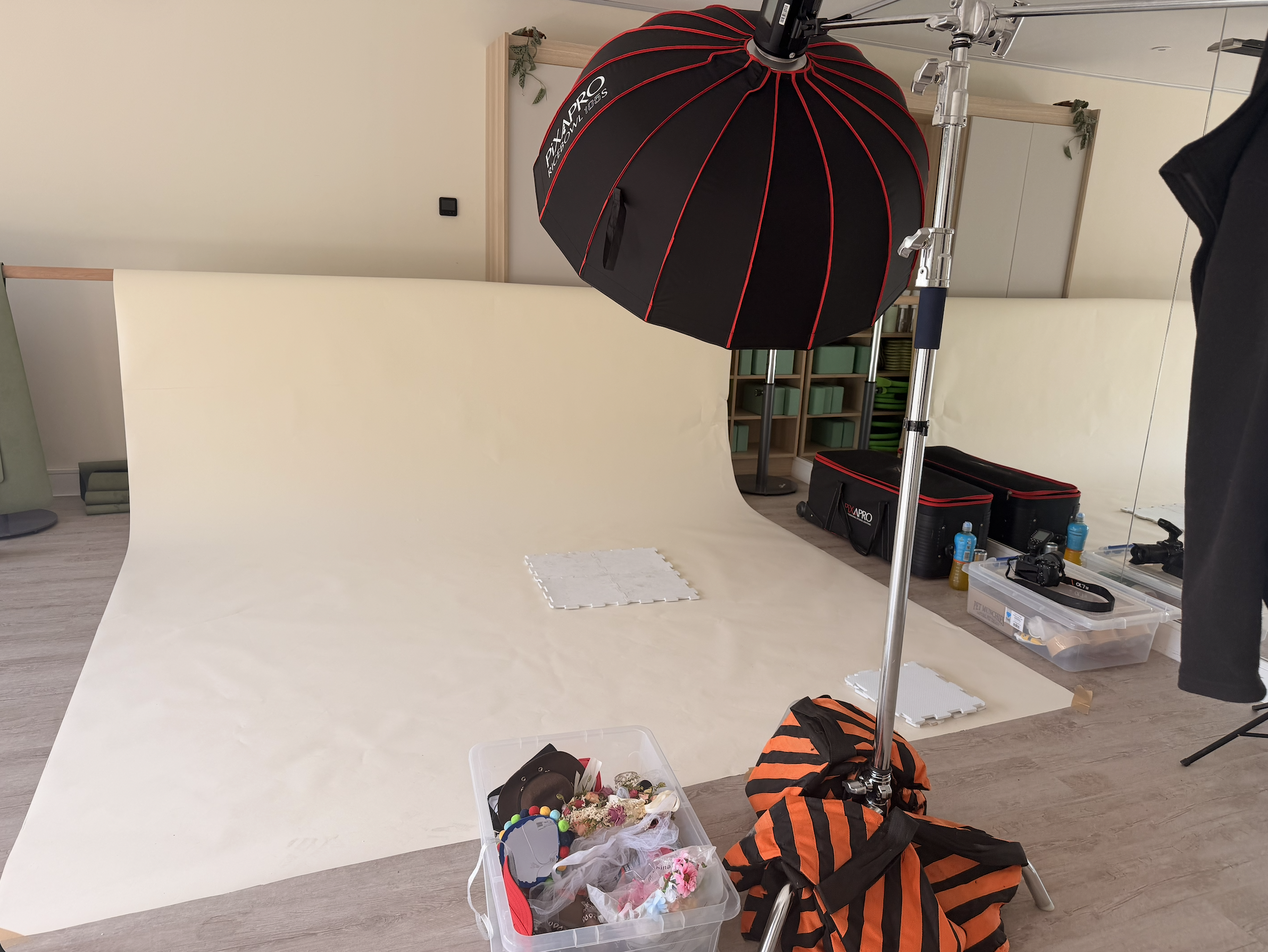 Photography studio setup with a white seamless backdrop, a large black and red photography umbrella, and various photography supplies including a bin of accessories and water bottles, with some furniture and storage in the background.