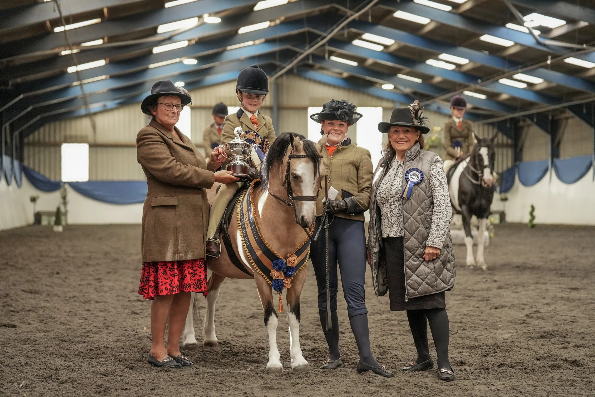 A group of people posing with a young girl riding a decorated horse inside an indoor equestrian arena. The girl is holding a trophy, and everyone is smiling and dressed in formal riding attire and hats.
