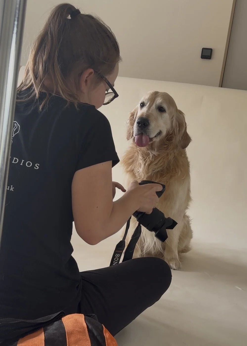 A woman with glasses holding a camera and kneeling next to a sitting golden retriever dog with its tongue out, in a room with plain walls.