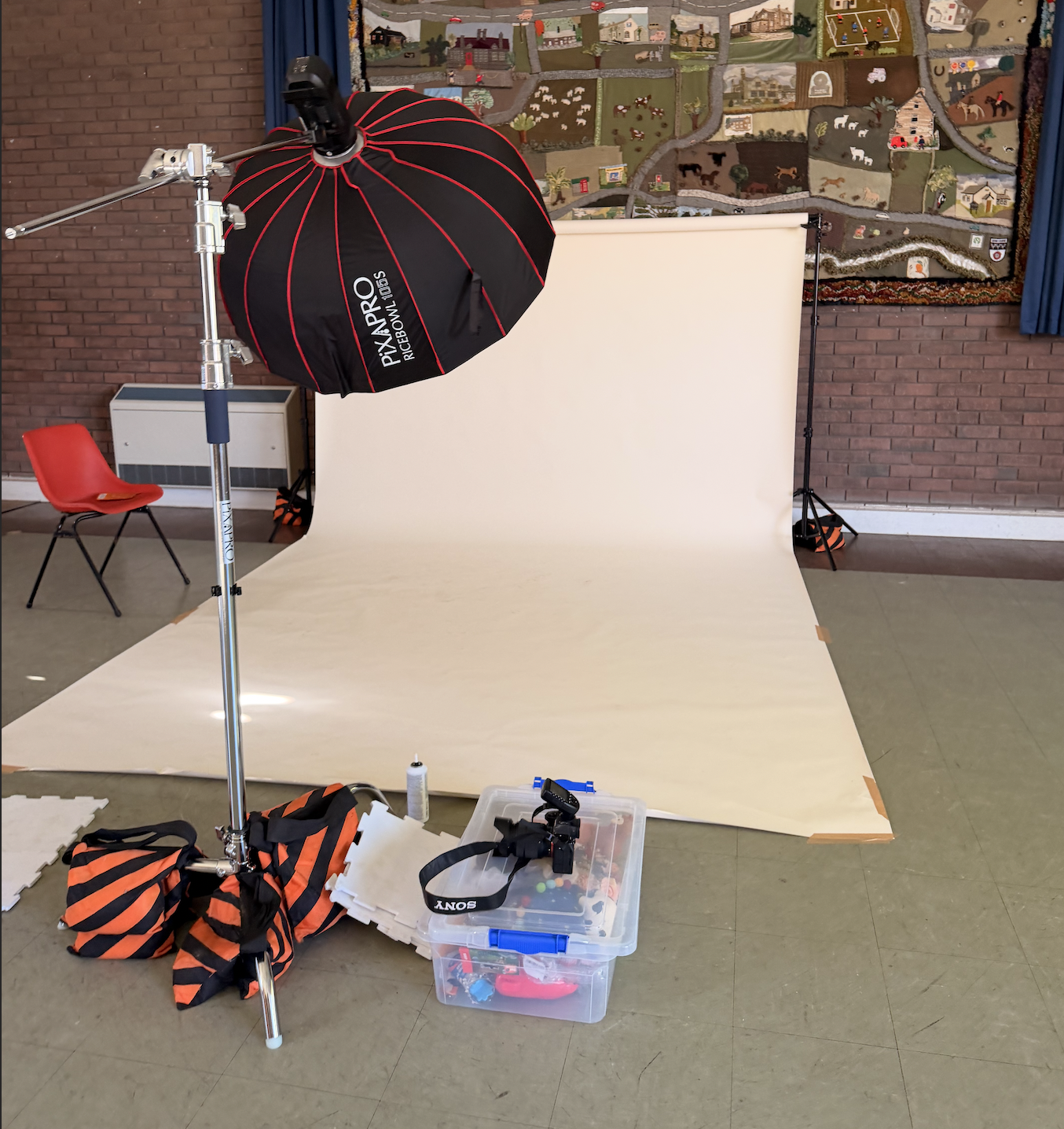 Photography setup with a white backdrop, studio light, camera, and equipment on the floor in front of a brick wall.