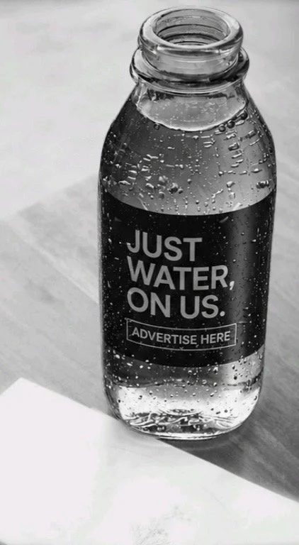 A glass water bottle filled with water, labeled 'Just Water, On Us' with a space for advertisement, placed on a table with a shadow cast on it.