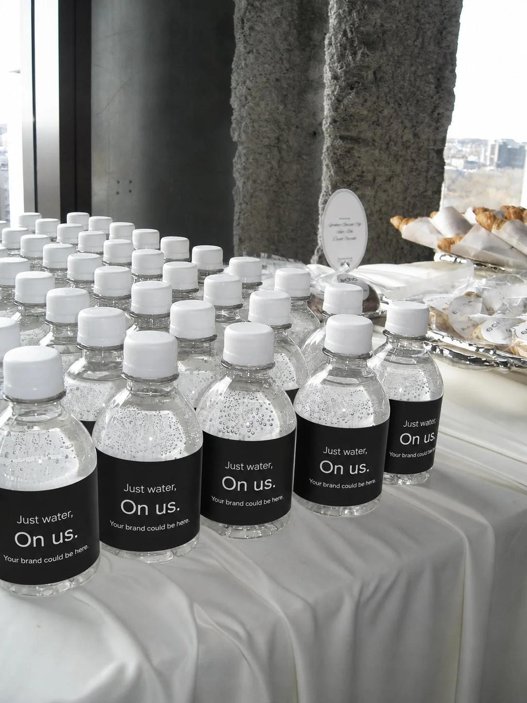 Bottles of water with black labels reading 'Just water, On us. Your brand could be here,' arranged on a table at an event or conference.