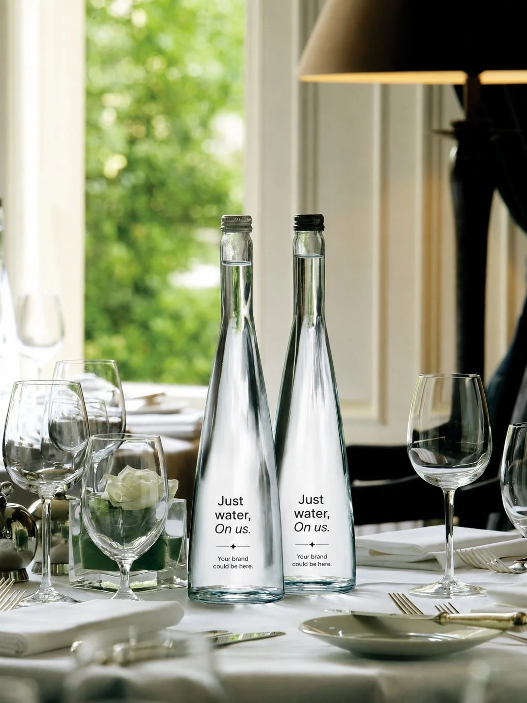 Two glass bottles labeled 'Just water, On us' placed on a dining table with wine glasses, plates, silverware, and a green background visible through a window.