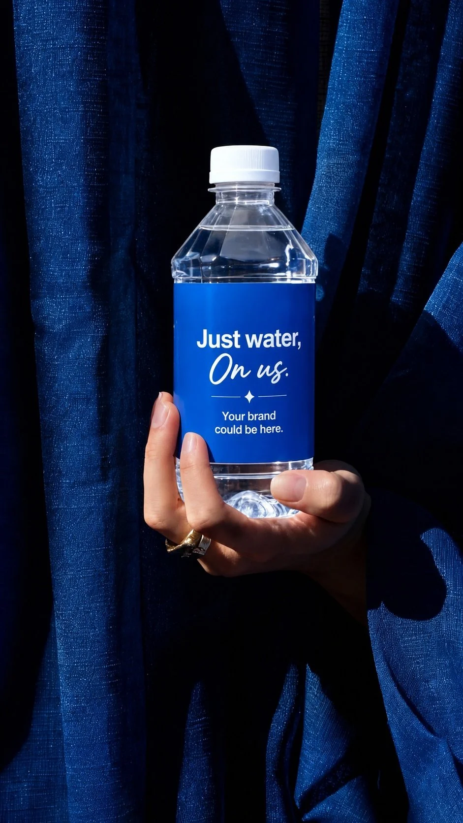 A hand holding a plastic water bottle with a blue label that reads 'Just water, On us. Your brand could be here.' in front of a dark blue textured curtain.