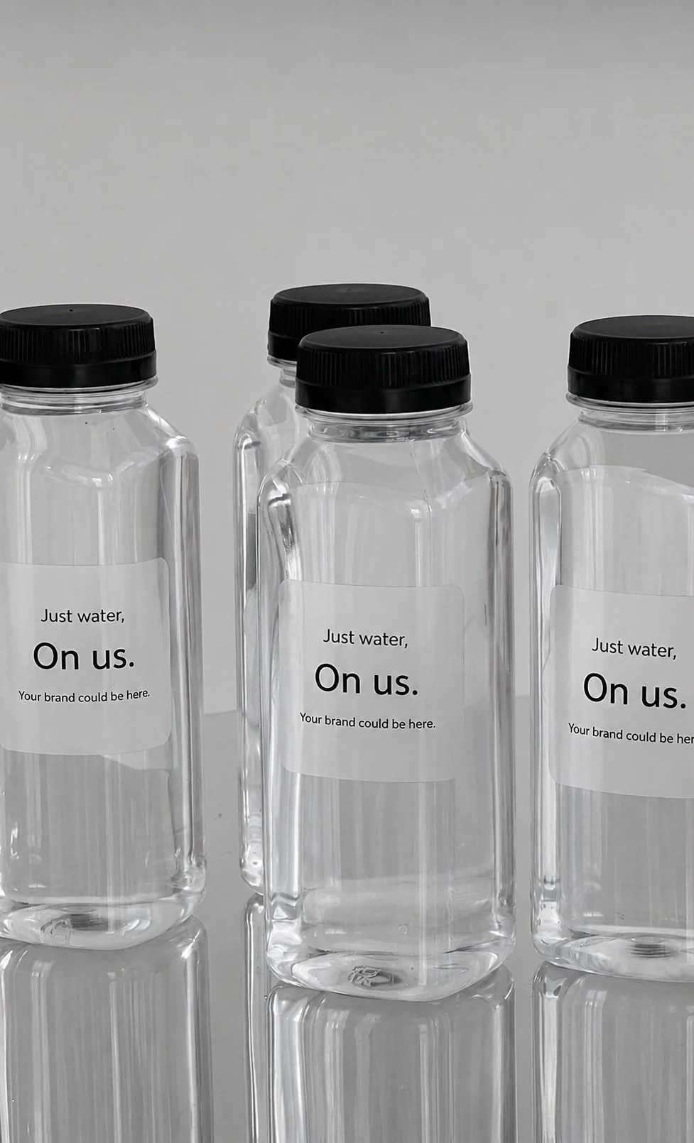 Four clear plastic water bottles with black caps, each with a blank label that says "Just water, On us. Your brand could be here." in black text, placed on a reflective surface against a plain gray background.